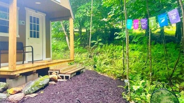 A small wooden cabin with a porch, surrounded by dense greenery and a string of colorful flags.