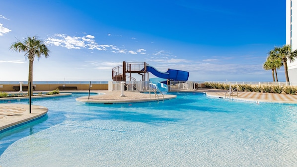 Una piscina con tobogán de agua, palmeras y un cielo azul despejado.