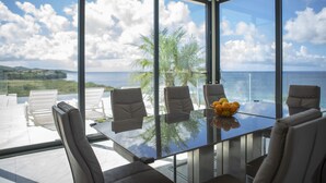 A modern dining area with a glass table and chairs, overlooking a scenic ocean view with palm trees and a clear sky. - Luxurious 6-Bedroom Villa with Private Pier & Panoramic Ocean View (Jeremi)