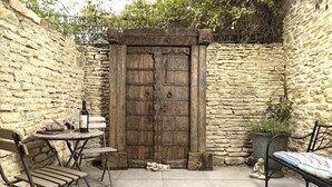 A stone-walled outdoor patio with a rustic wooden door, a small round table with a bottle, and two metal chairs.