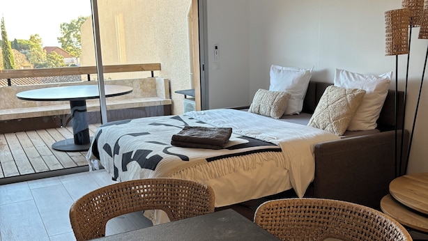 Een hotelkamer met een bed, een eettafel met stoelen, een balkon met een tafel en stoelen, en een groot raam.