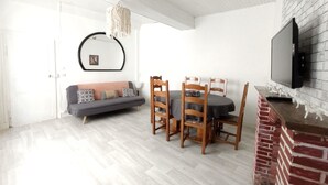 A dining area with a table and chairs, a sofa, a television mounted on a brick wall, and a hanging light fixture.