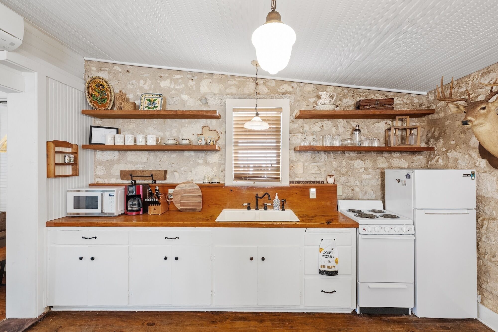 A rustic kitchen with a stone wall, wooden shelves, a white refrigerator, a stove, and a wooden cabinet with drawers.