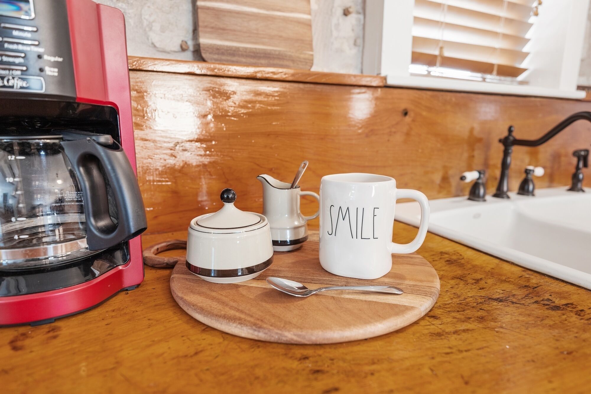 A kitchen counter with a coffee maker, a mug with "SMILE" written on it, a small pitcher, and a spoon.