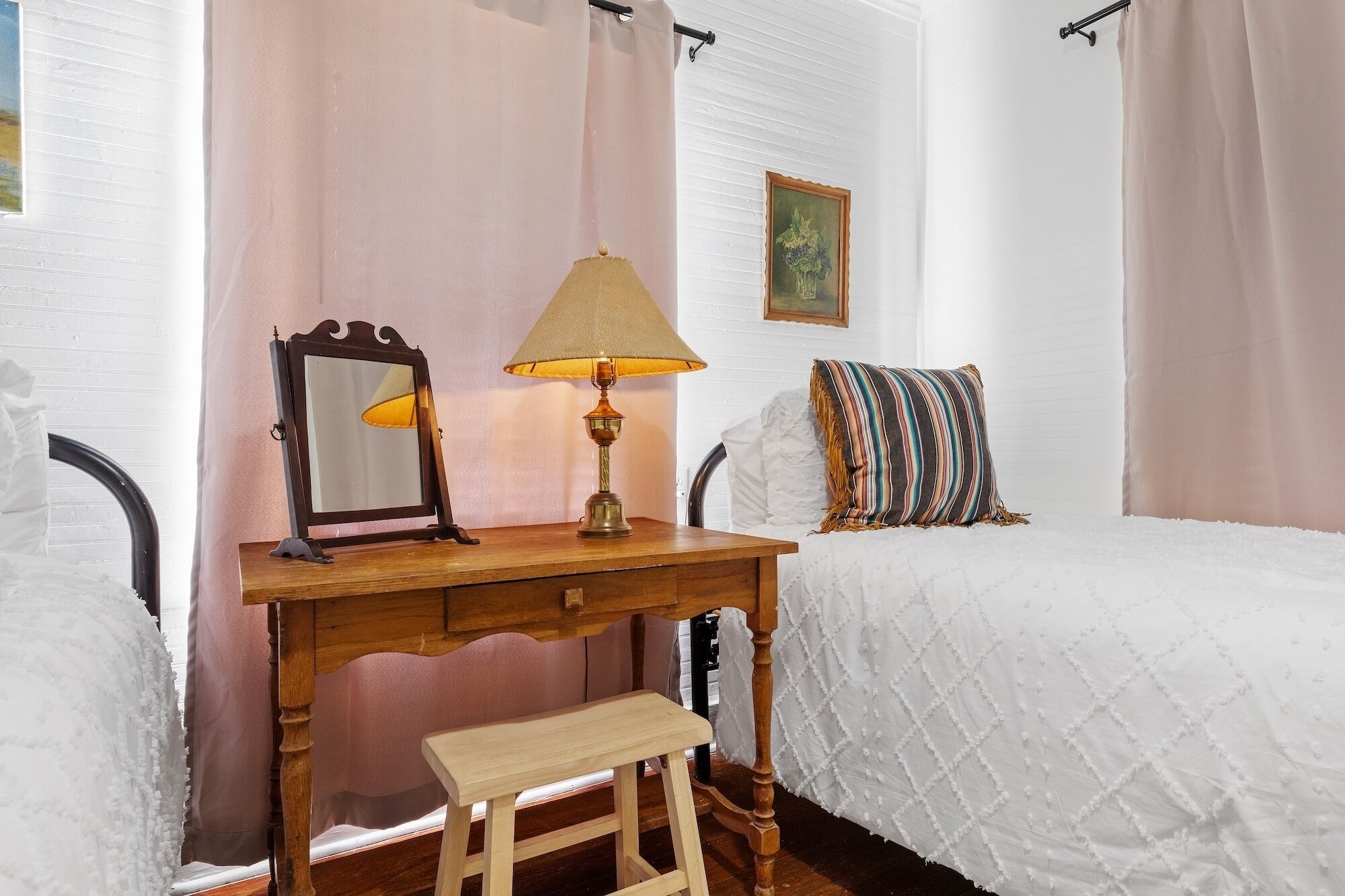A bedroom with a wooden desk, a small stool, a bed with a white quilt, a striped pillow, a lamp, and a framed mirror.