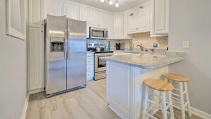 A modern kitchen with white cabinetry, a stainless steel refrigerator, a microwave, and a granite countertop with two wooden stools. - Oceanfront North Topsail Beach Condo w/Pool! Linens and towels provided (North Topsail Beach)