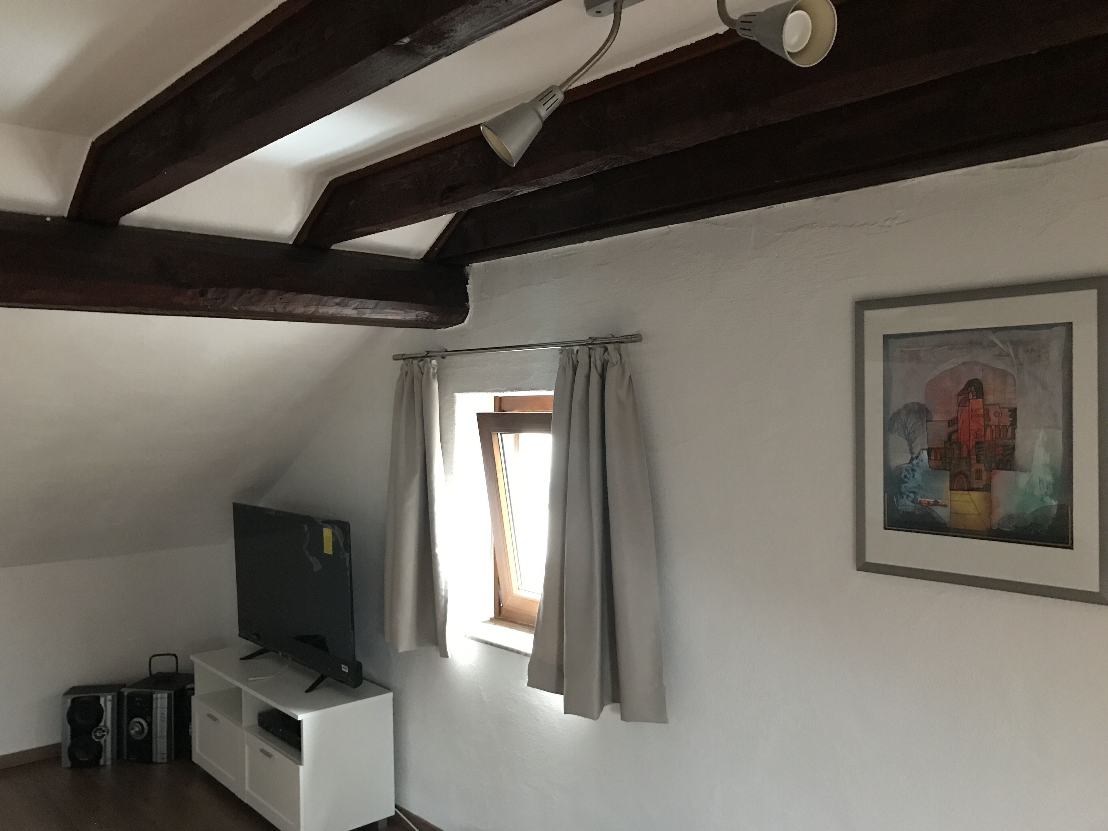 Una habitación con una televisión sobre un estante blanco, un cuadro en la pared y una ventana con cortinas.