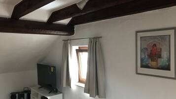 Una habitación con una televisión sobre un estante blanco, un cuadro en la pared y una ventana con cortinas.
