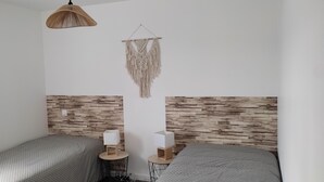 Habitació amb dos llits, ambures de fusta, un penjoll de macramè, una làmpada penjant de fibra natural i una taula rodona petita amb una làmpada.