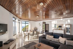 Un salon moderne avec un plafond en bois, un canapé gris, un coin repas et un téléviseur fixé au mur.