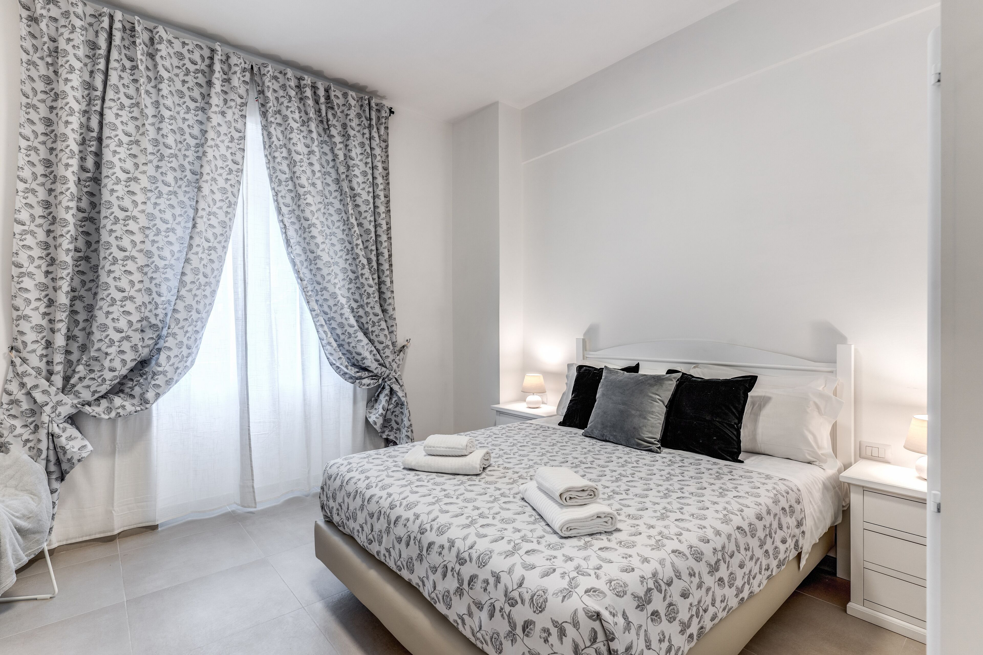 Ein modernes Schlafzimmer mit einem großen Bett, weißen Vorhängen und einem Nachttisch mit einer Lampe.
