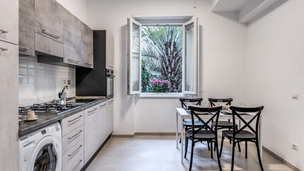 A compact kitchen with a built-in washer, a dining area with four chairs, and a window with a view of greenery. - PMT APT (Rome)