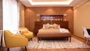 A modern hotel room with a large bed, a cityscape painting on the wall, a small round table, and two yellow armchairs.