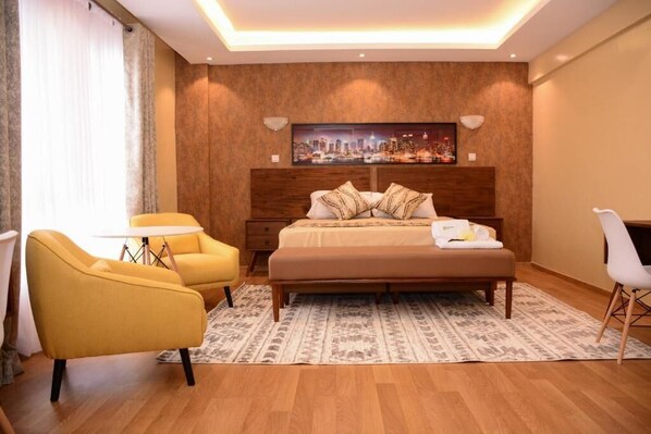 A modern hotel room with a large bed, a cityscape painting on the wall, a small round table, and two yellow armchairs. - Lux Suites Westlands Penthouse (Nairobi)