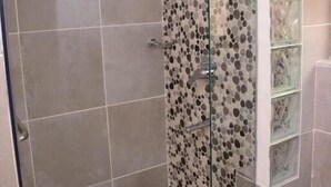 A shower with a glass door, tiled walls, and a glass shelf unit.