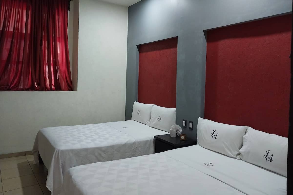 A hotel room with two beds, each with white bedding and pillows marked with initials. The room has a red curtain, a small table with a lamp, and a wall-mounted light fixture.