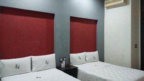 Habitación de hotel con dos camas, cada una con ropa de cama y almohadas blancas, cabeceros rojos y una mesita de noche pequeña con una lámpara y una caja de pañuelos.