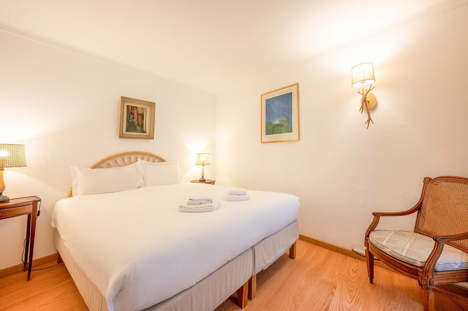 A hotel room with a large bed, two bedside lamps, a chair, and two framed pictures on the wall.