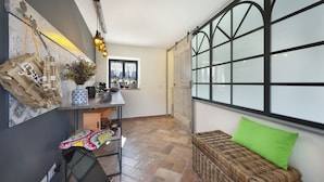 A cozy room with a wicker sofa, a small table with decorative items, a window with black frames, and a wall-mounted light fixture. - YourHome - Karme House (Meta)