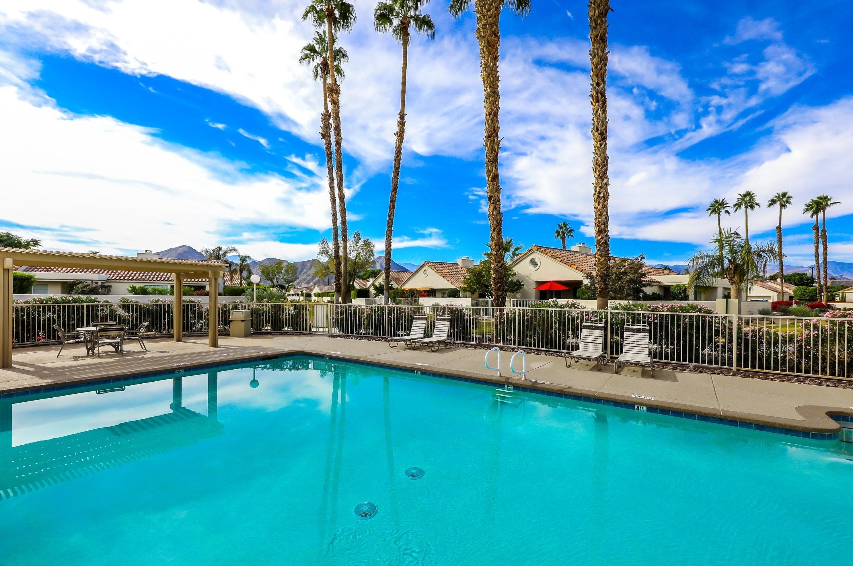 A swimming pool with lounge chairs and a pergola, surrounded by palm trees and a clear sky.