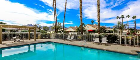 A swimming pool with lounge chairs and a pergola, surrounded by palm trees and a clear sky.