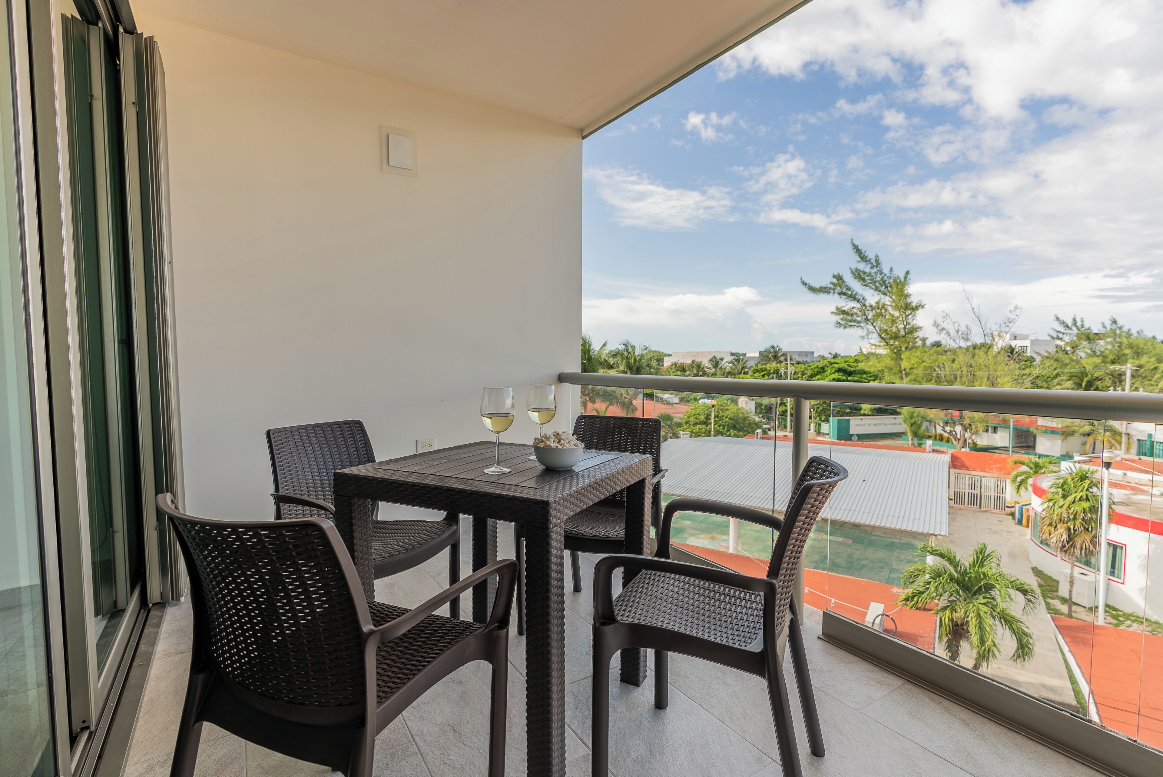Un balcón con mesa y sillas, un ventanal de vidrio y vistas a edificios y árboles.