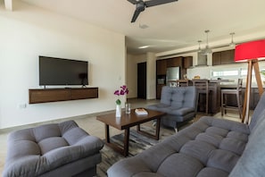 A modern living room with a flat-screen TV, grey sofas, a coffee table, and a kitchen area in the background. - La Poza (Isla Mujeres)