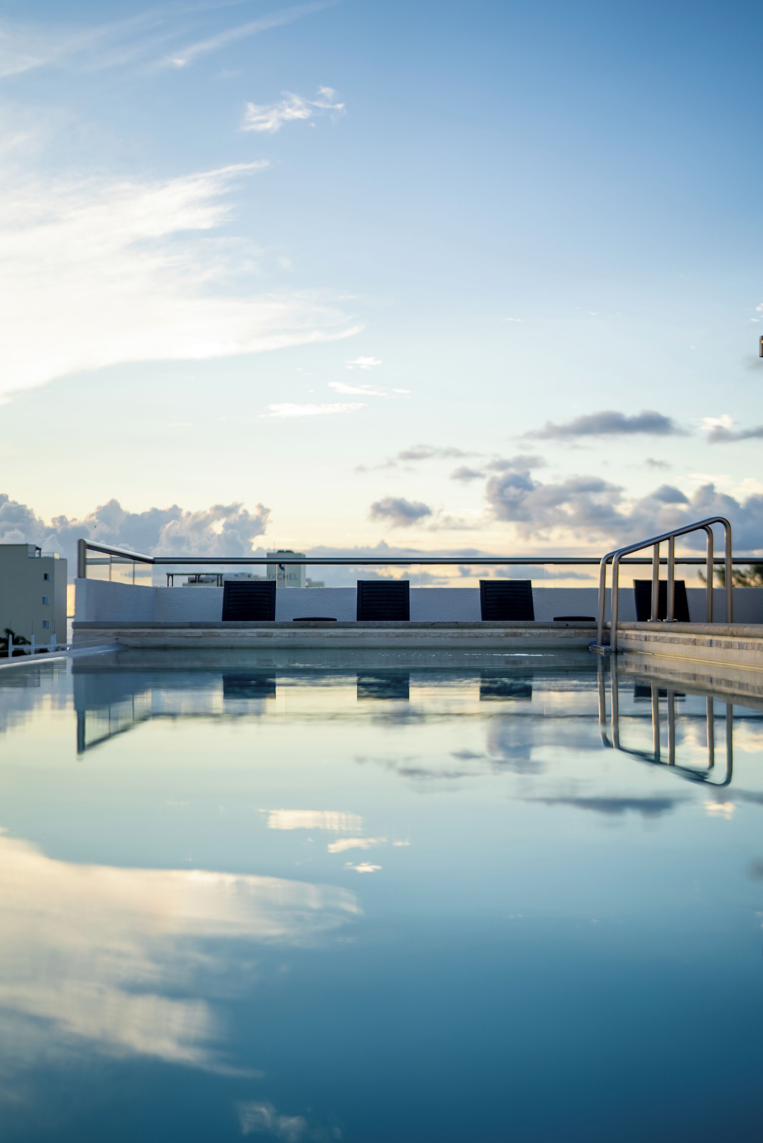 Una piscina con una escalera de metal, un muro blanco y un cielo despejado con nubes.