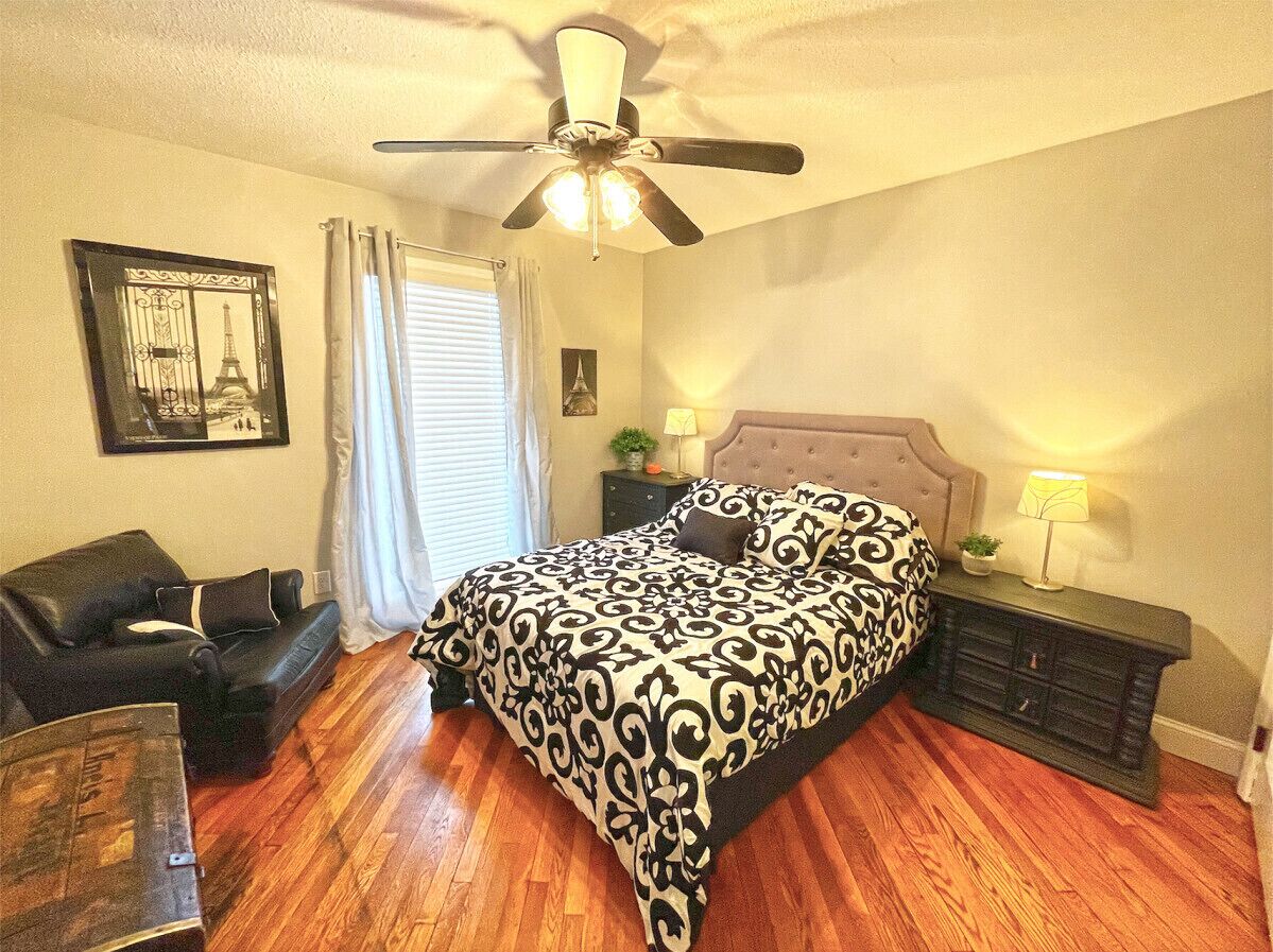 Une chambre à coucher avec un lit, un ventilateur de plafond, une table de chevet, une lampe, une fenêtre avec des rideaux, un tableau encadré au mur et un plancher en bois.