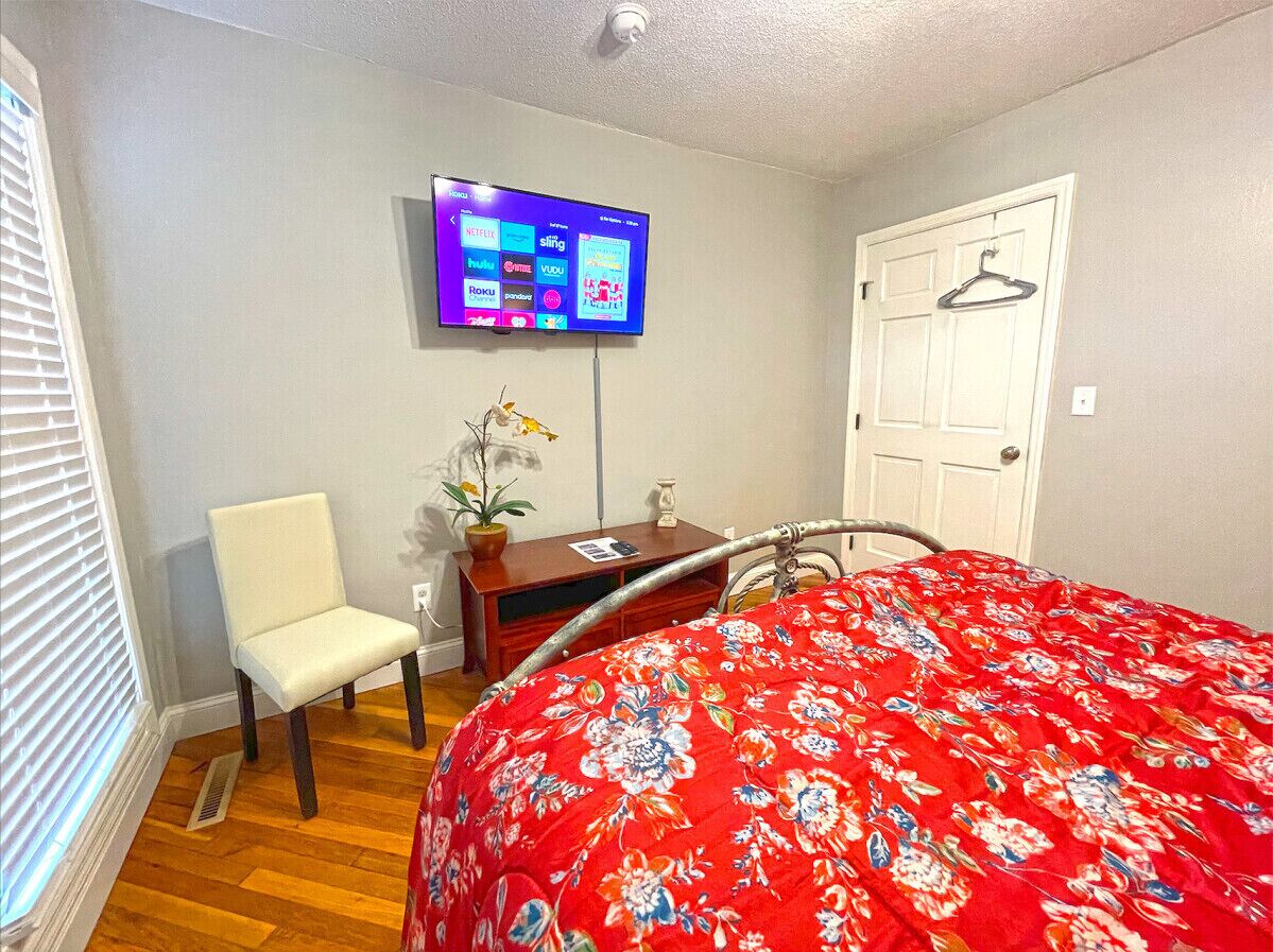 A bedroom with a bed, a chair, a desk, a TV, and a door.