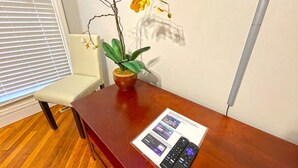 Ein Holztisch mit einer Fernbedienung und einer Broschüre, einer Topf-Orchidee und einem Stuhl in einem Zimmer mit Holzboden und einem Fenster mit Jalousien.