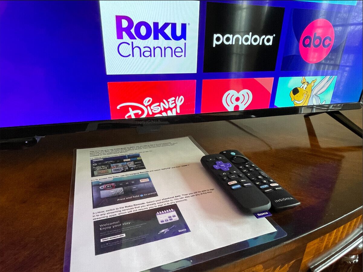 A TV screen displaying Roku Channel, Pandora, and ABC logos, a remote control, and a printed guide on a glass table.