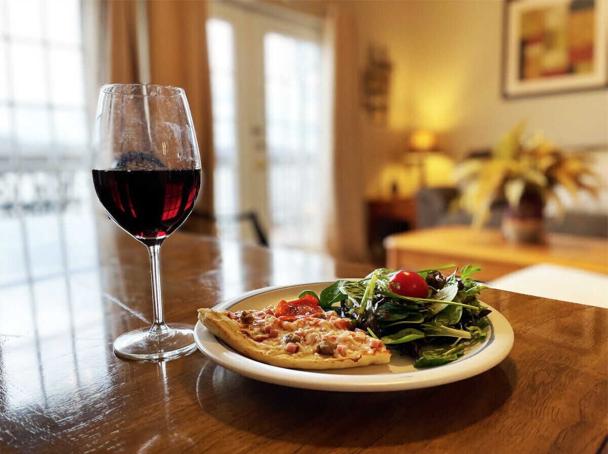 A dining table with a plate of pizza and a glass of red wine, and a window with a view of the outdoors.