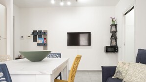 Ett modernt vardagsrum med ett vitt matbord, en stor grön skål, en gul stol, en mörk soffa och en platt-TV som är monterad på väggen.