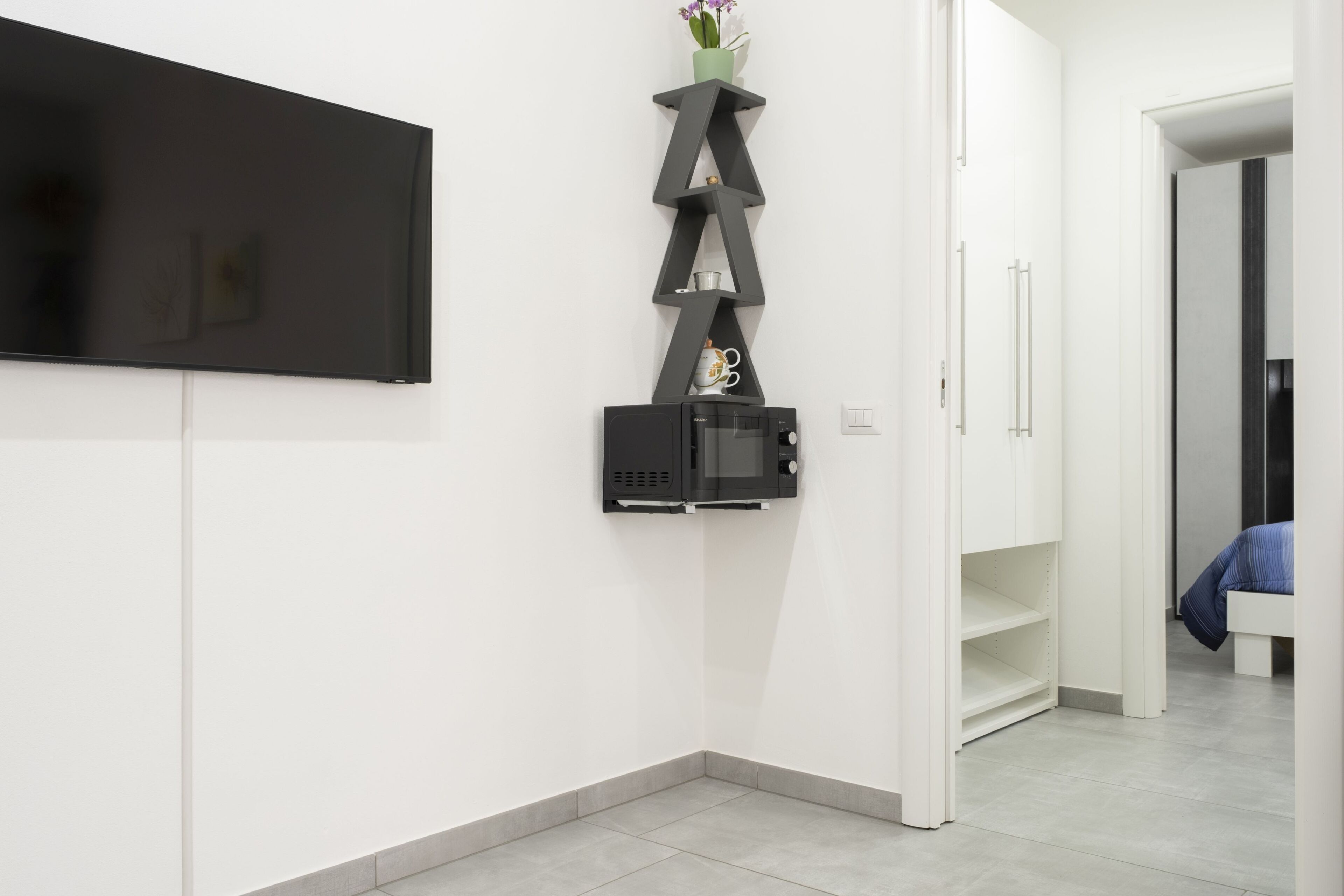 A modern room with a flat-screen TV mounted on the wall, a built-in microwave, and a shelving unit with a vase and mug.