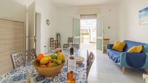 A dining area with a table set for two, a bowl of fruit, and a bottle of liquor. There is a blue sofa with yellow cushions in the background. - Villetta Punta Grossa short walk to beach (Porto Cesareo)