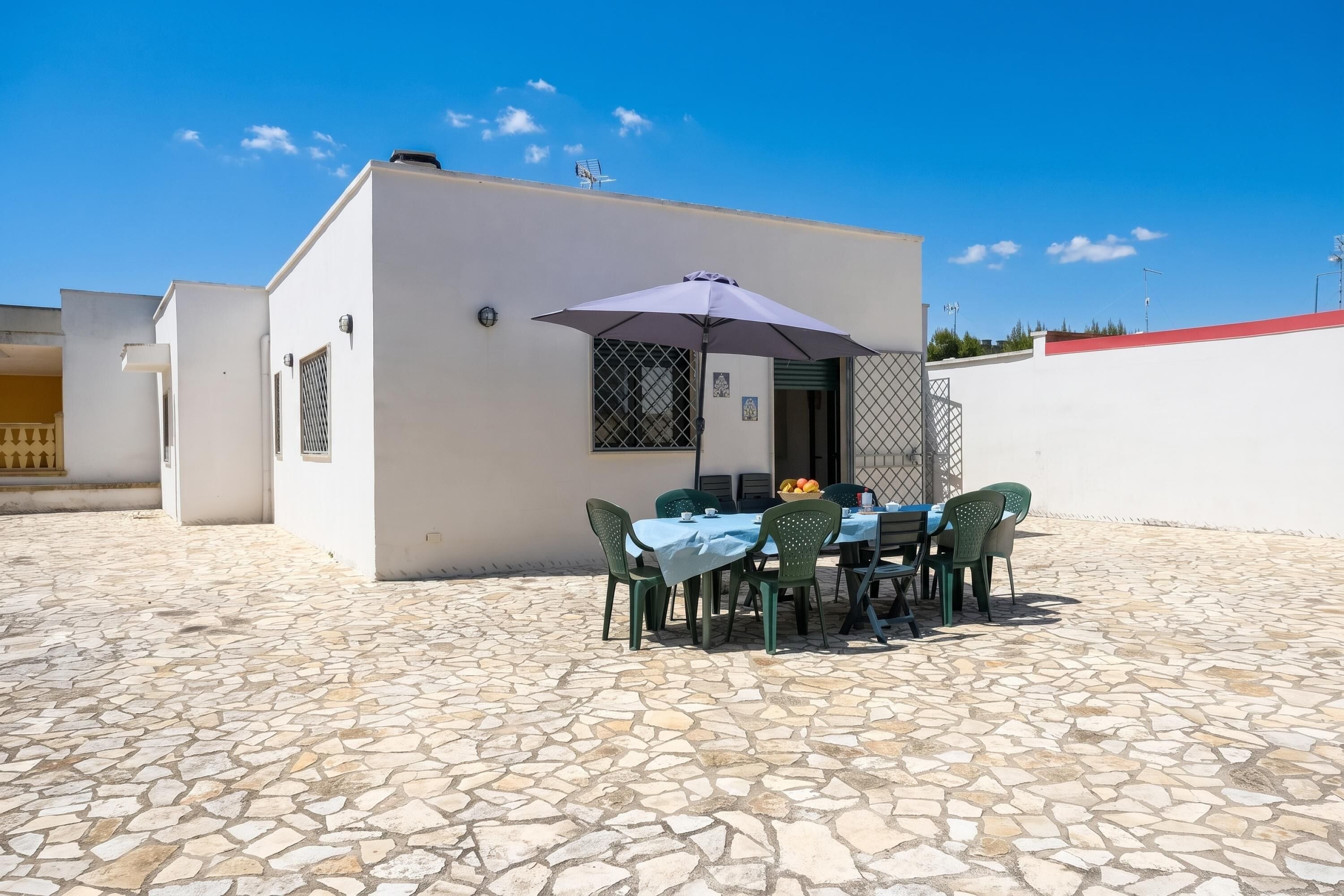 Un patio con una mesa y sillas, una sombrilla cubierta y un edificio blanco de techo rojo.