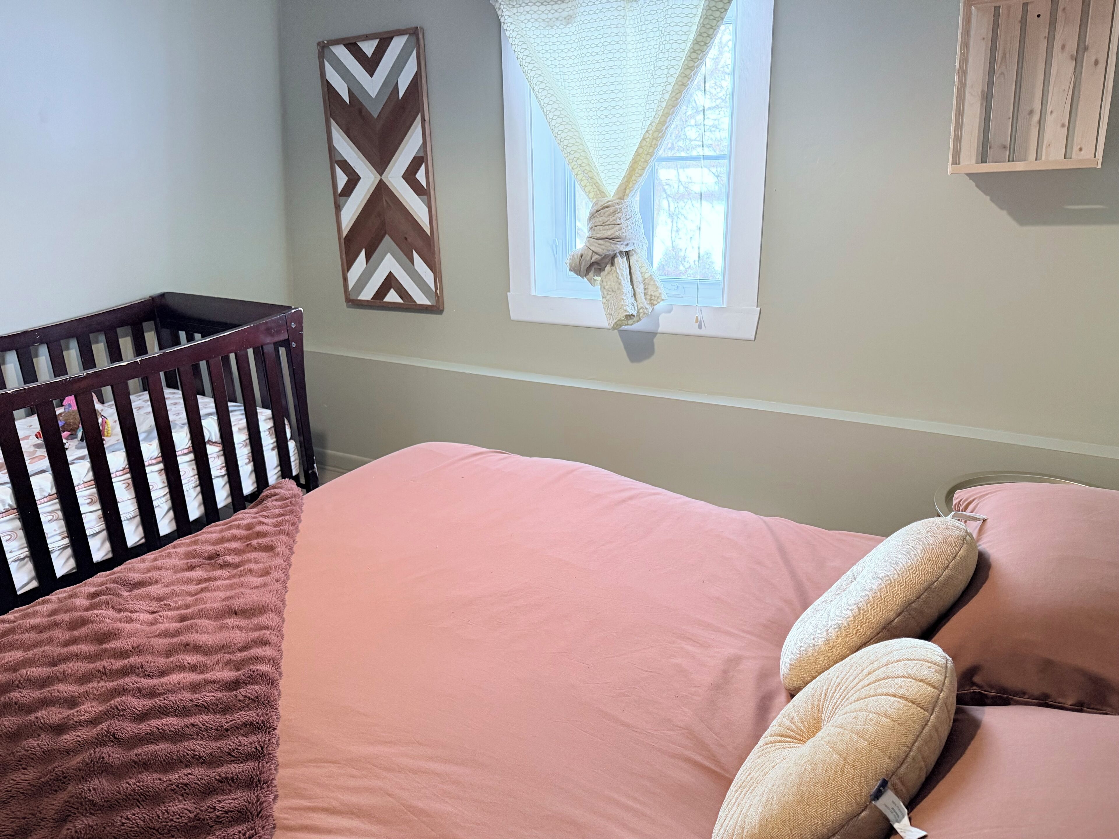 A baby crib with a patterned mattress, a pink bed with two pillows, a decorative wall piece, and a window with a white curtain.