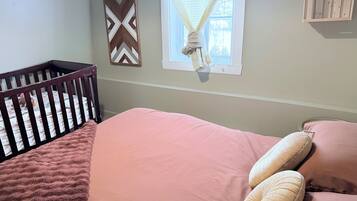 A baby crib with a patterned mattress, a pink bed with two pillows, a decorative wall piece, and a window with a white curtain.