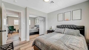 Een slaapkamer met een groot bed, een nachtkastje en een stoel. Een deur leidt naar een andere kamer.
