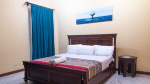 A neatly made bed with a colorful blanket and a rolled towel, a wooden nightstand beside it, and a painting of a whale on the wall. - Lux Suites Le Royal Apartments Nyali (Mombasa)
