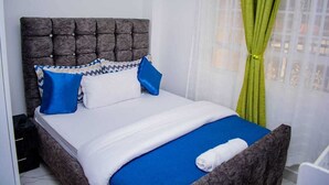 Una cama con cabecera capitonada, ropa de cama blanca, almohadas azules y blancas, y una cortina verde.