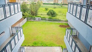 Vista desde un balcón sobre un área verde con cercado, árboles y edificios a lo lejos.