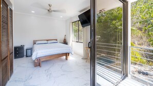 A bedroom with a bed, a ceiling fan, a TV, and a balcony with a view of trees.