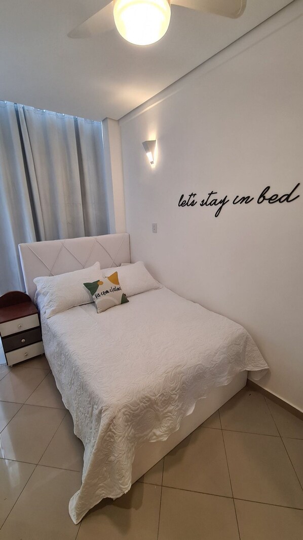 A neatly made bed with a white bedspread and two pillows, one with a Christmas-themed design. A wall-mounted light fixture and a wall with the text "let's stay in bed" are visible.