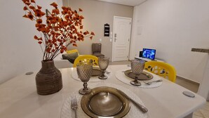 Comedor con mesa dispuesta para cuatro, que incluye un jarrón de flores naranjas, cristalería y un televisor mostrando un programa.