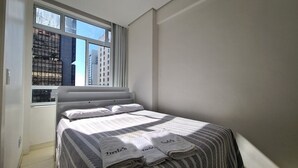 Una cama bien hecha con ropa de cama y toallas a rayas, una ventana con vistas a edificios y una pared lisa.