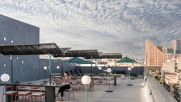 An outdoor dining area with tables and chairs, umbrellas, and a cityscape in the background.