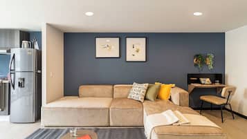 Una sala de estar moderna con un sofá modular, una mesa de centro y una mesita auxiliar. Se ve un refrigerador y una zona de cocina a la izquierda.