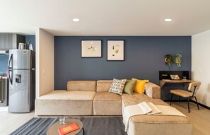 A modern living room with a sectional sofa, a coffee table, and a small side table. There is a refrigerator and a kitchen area visible to the left. - Kukun Sócrates Polanco (Mexico City)